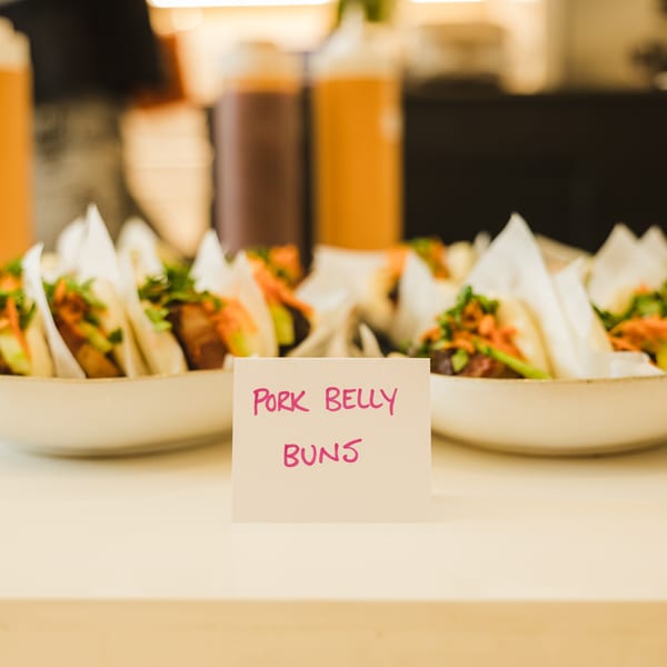 Plates of pork belly buns with vegetables and sauces on a counter, with a "Pork Belly Buns" sign.