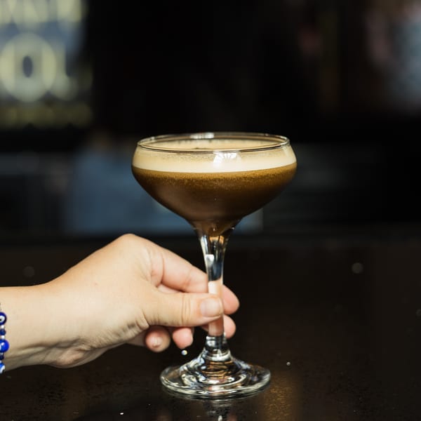 Hand holding a coupe glass filled with a dark, foamy espresso martini on a dark bar.