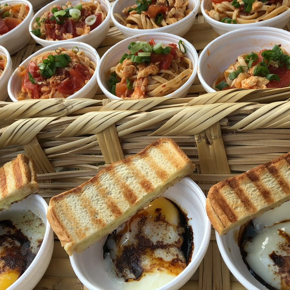 A bamboo tray holds small bowls of pasta with tomatoes and green onions, and soft-boiled eggs with toast.