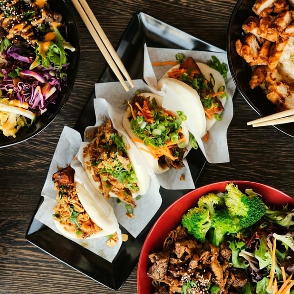 An overhead shot of various Asian dishes, including shrimp, chicken, and beef bowls, and bao buns.