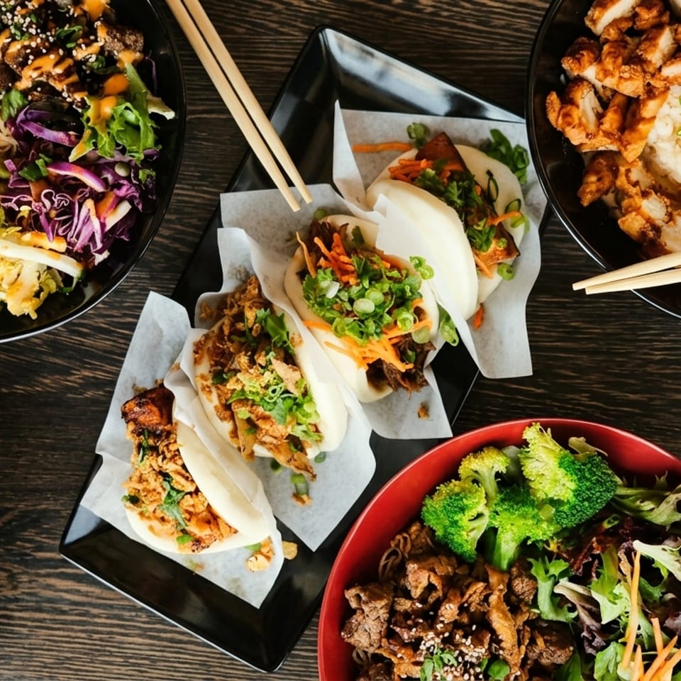 An overhead shot of various Asian dishes, including shrimp, chicken, and beef bowls, and bao buns.