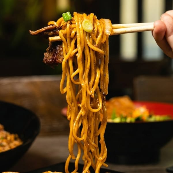 Chopsticks lift a tangle of golden noodles with beef and green onions from a bowl.