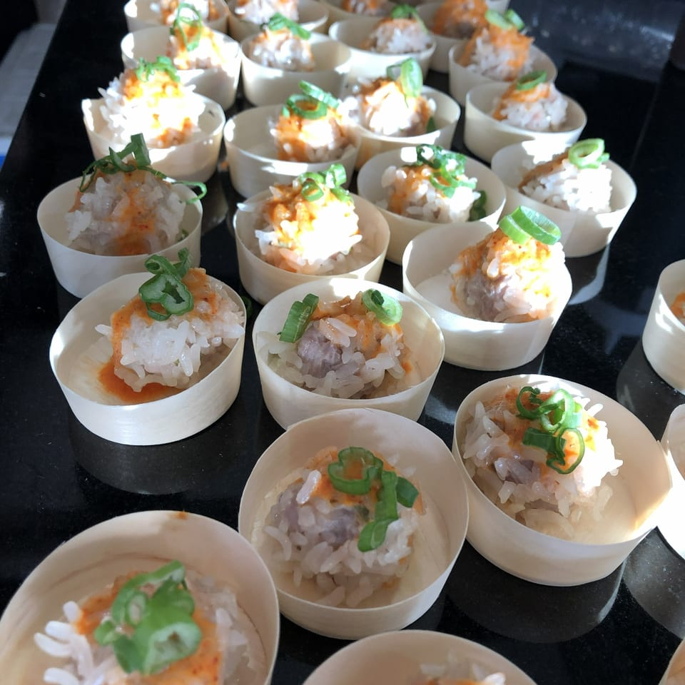 Rows of small wooden cups filled with rice, meat, orange sauce, and green onions.