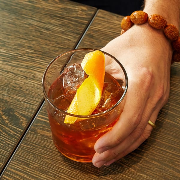 A hand holding a cocktail with ice and an orange peel garnish on a wooden table.