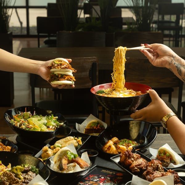 Hands holding a stack of bao buns and chopsticks with ramen, surrounded by various Asian dishes.