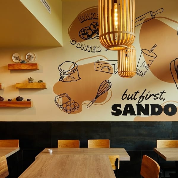 Restaurant interior with light wood tables, chairs, and a wall mural depicting baking ingredients and boba.