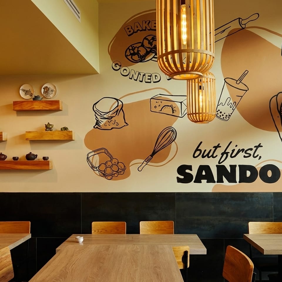 Warmly lit restaurant dining area with wooden tables, shelves of teapots, hanging lights, and a mural reading “Boba is life” and “Sando.”