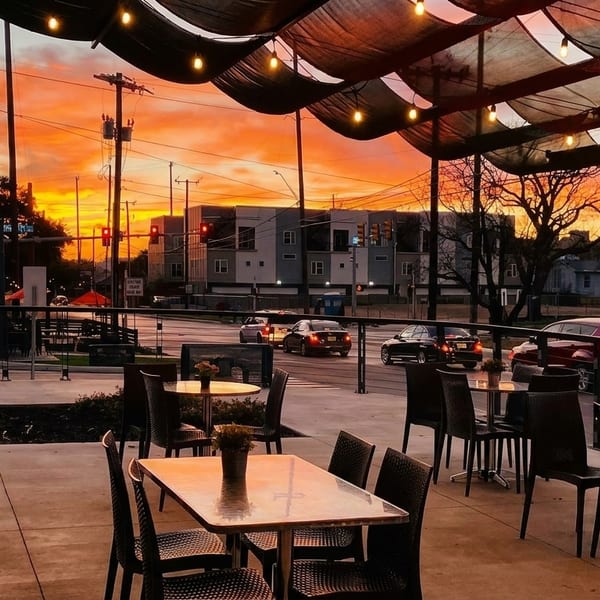 Restaurant patio with empty tables and chairs under string lights, overlooking a street at sunset.