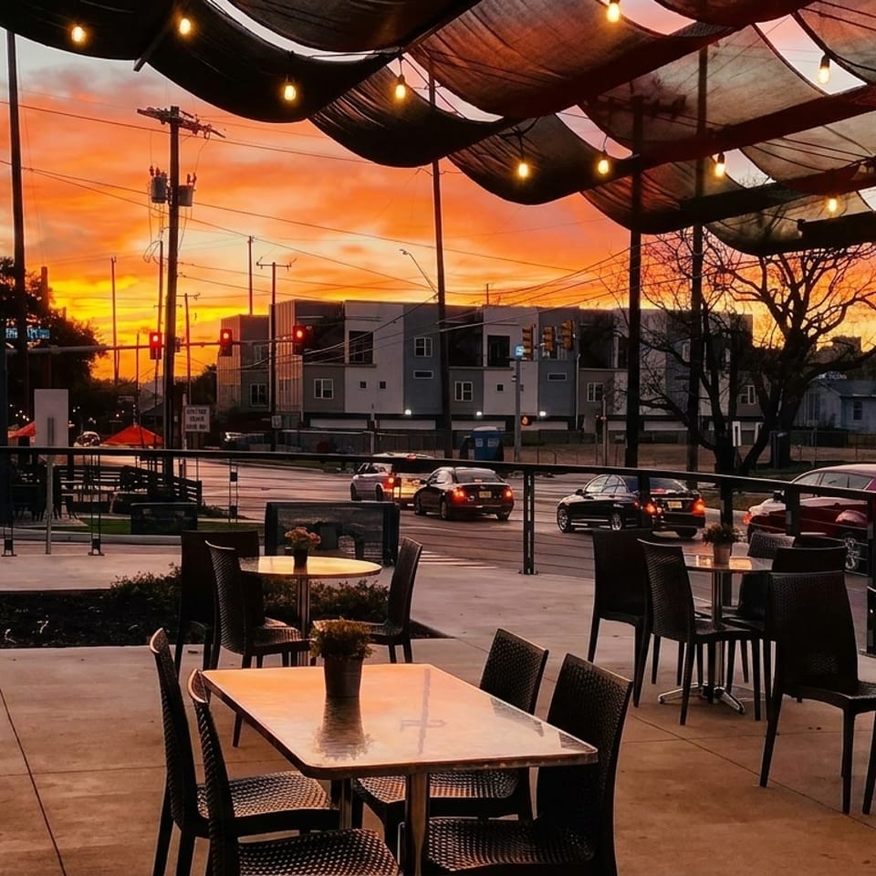 Restaurant patio with empty tables and chairs under string lights, overlooking a street at sunset.