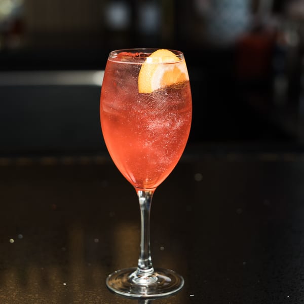 A vibrant red cocktail with an orange slice garnish in a wine glass on a dark, reflective surface.
