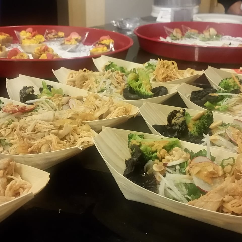 Rows of small wooden boats filled with Asian noodles, shredded chicken, broccoli, and mushrooms.