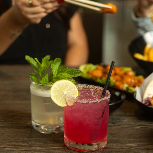 Two cocktails on a wooden table, with a person using chopsticks to pick up food in the background.