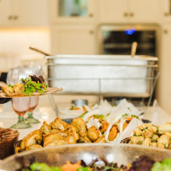 A gloved hand serves a plate of dumplings and salad from a buffet spread with various appetizers.