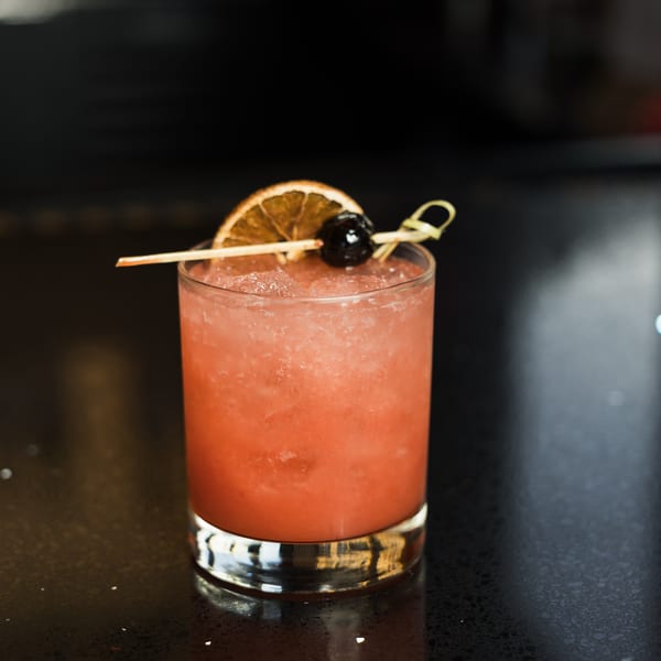 A vibrant pink cocktail, garnished with a dried orange slice and a dark cherry, sits on a dark surface.
