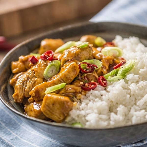A bowl of chicken stir-fry with white rice, garnished with sliced red chilies and green onions.