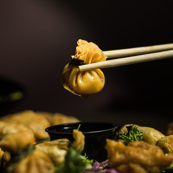 Chopsticks holding a golden fried dumpling, dripping sauce, above a plate of appetizers.