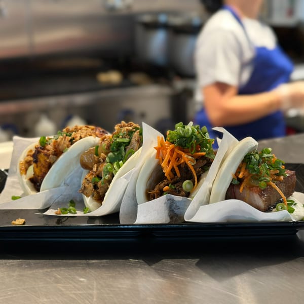 A black rectangular plate holds five gourmet bao buns with various fillings, on a metal counter.