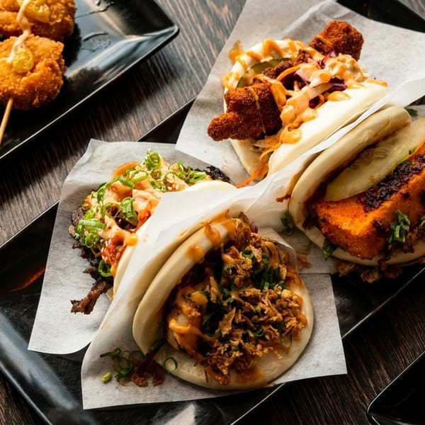 Assortment of bao buns with various fillings, fried shrimp skewers, and beef skewers on black plates.