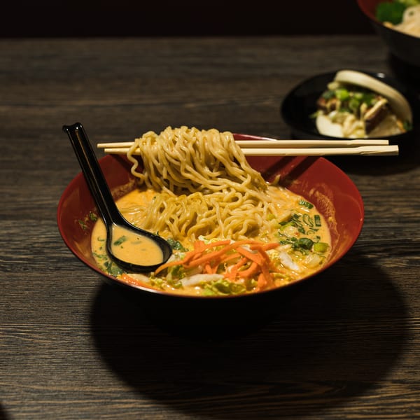 A red bowl of ramen with chopsticks and a spoon, alongside a cocktail and a bao bun.