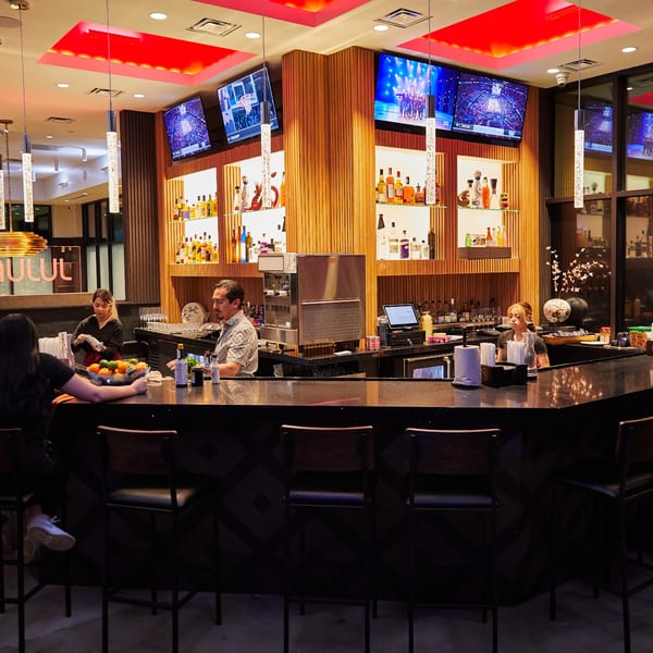 A modern restaurant bar with patrons, bartenders, liquor shelves, and multiple TVs showing sports.