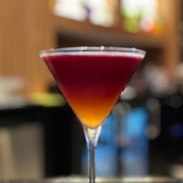 A vibrant, layered cocktail in a martini glass sits on a bar mat, with a blurred background.