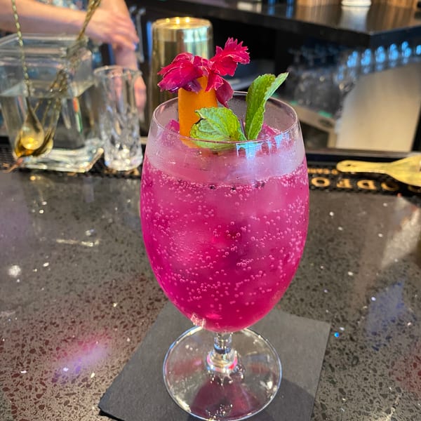 A vibrant pink cocktail, garnished with orange peel, mint, and a pink flower, sits on a bar.