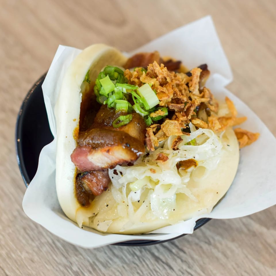 A bao bun filled with pork belly, sauce, fried onions, and green onions on a wooden table.