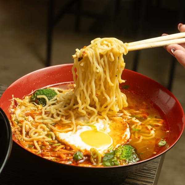 Chopsticks lifting noodles from a red bowl of ramen with egg, broccoli, and broth.