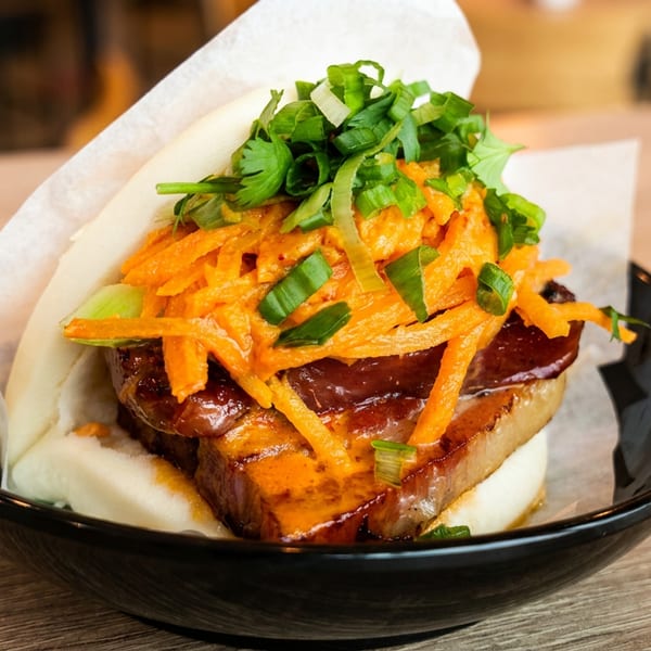 Pork belly bao bun with shredded carrots, green onions, and cilantro in a black bowl.