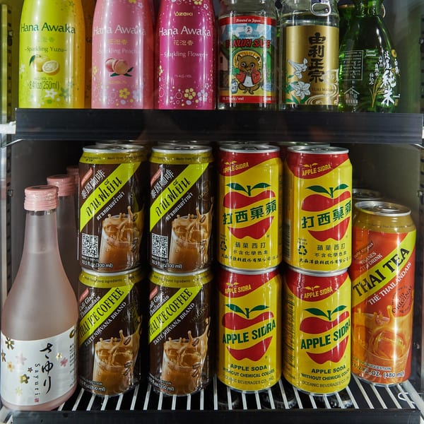 A refrigerated display case filled with various Asian beverages, including sparkling sake, iced coffee, apple soda, and Thai tea.