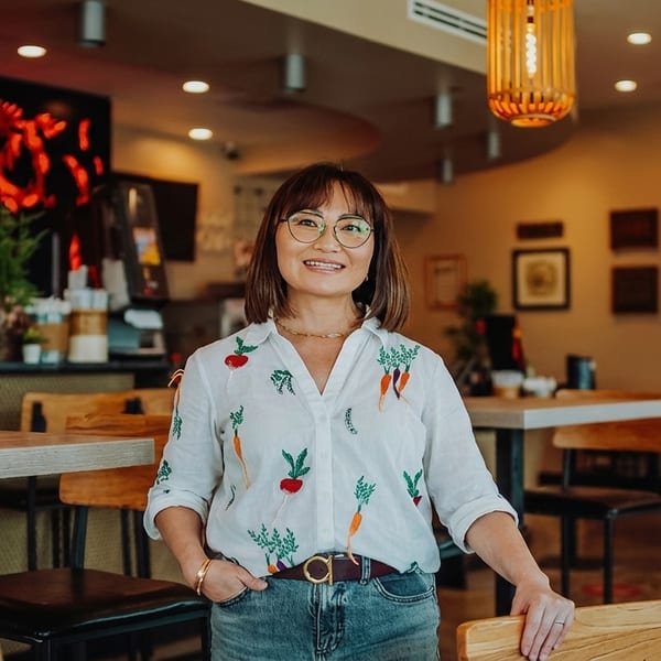 Smiling woman in a vegetable-patterned shirt stands in a modern restaurant dining area.
