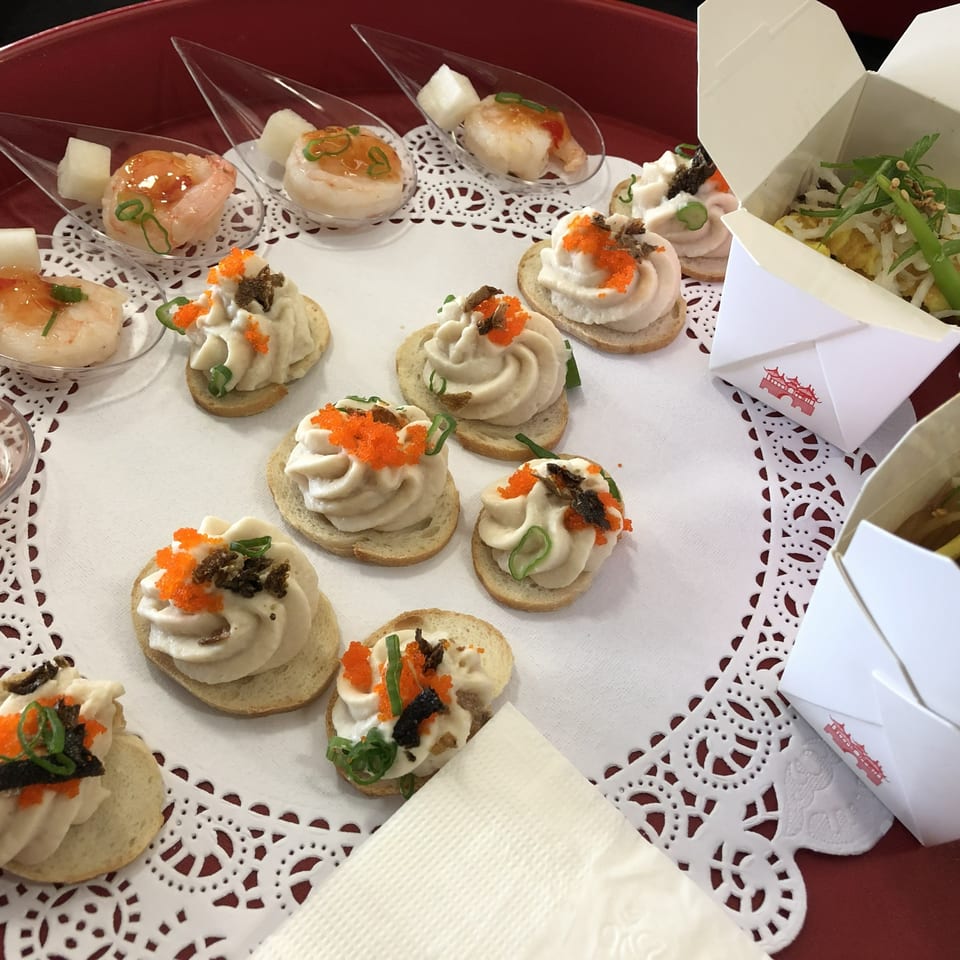 A red tray holds various appetizers, including shrimp in spoons, canapés, and small noodle boxes.