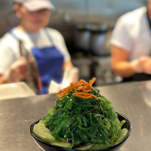 A vibrant green seaweed salad with cucumber and shredded carrots in a black bowl on a metal counter.