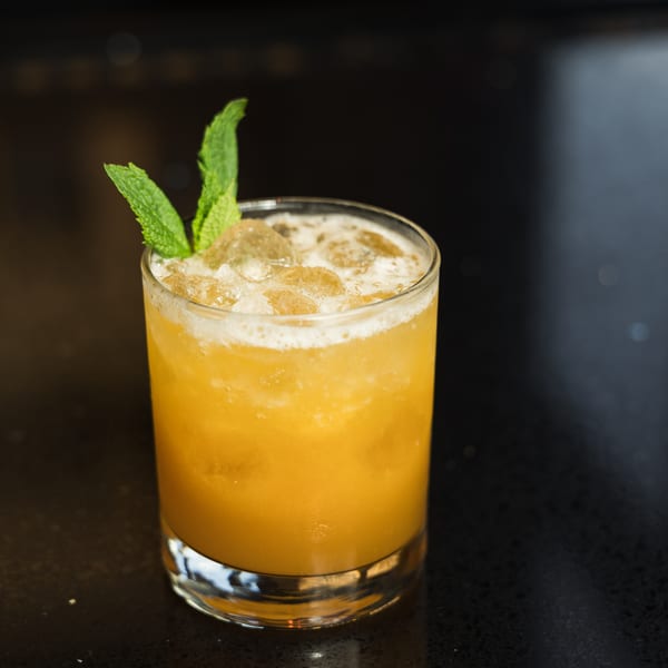 A vibrant orange cocktail with ice and a mint garnish sits on a dark, reflective surface.