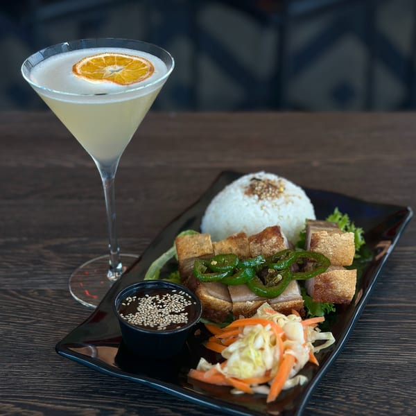 Crispy pork belly with rice, pickled vegetables, and a cocktail garnished with an orange slice.