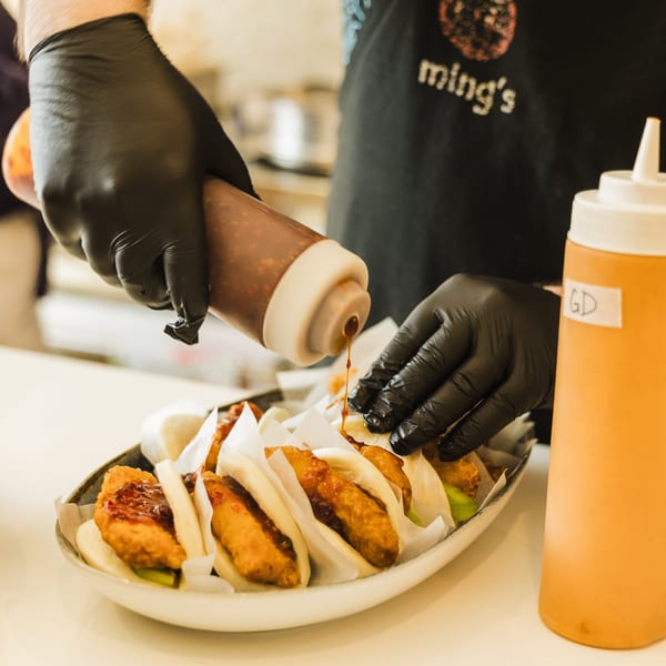 Chef in black gloves drizzles sauce onto a plate of Ming's fried chicken bao buns.