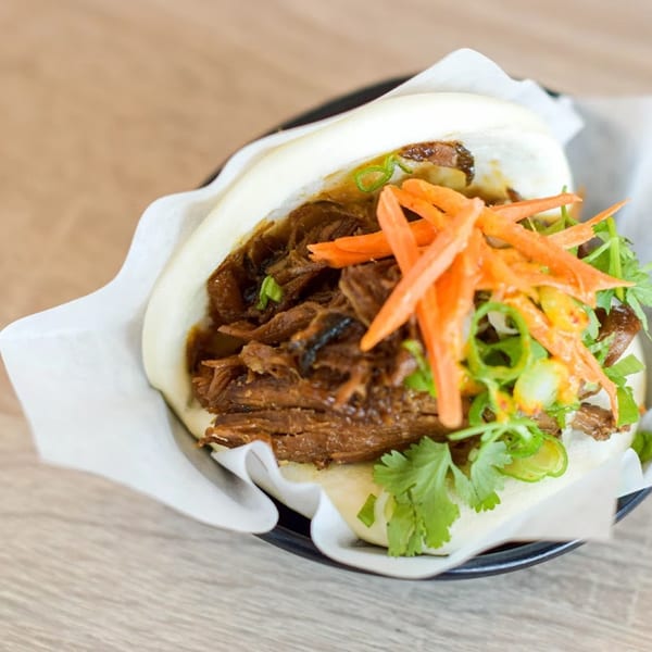 A fluffy bao bun filled with braised meat, shredded carrots, cilantro, and green onions.