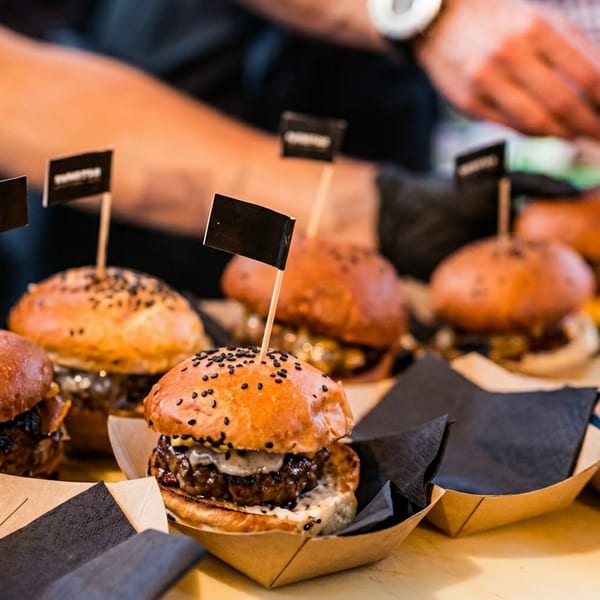 Mini burgers with black sesame buns and flags, served in individual paper trays.