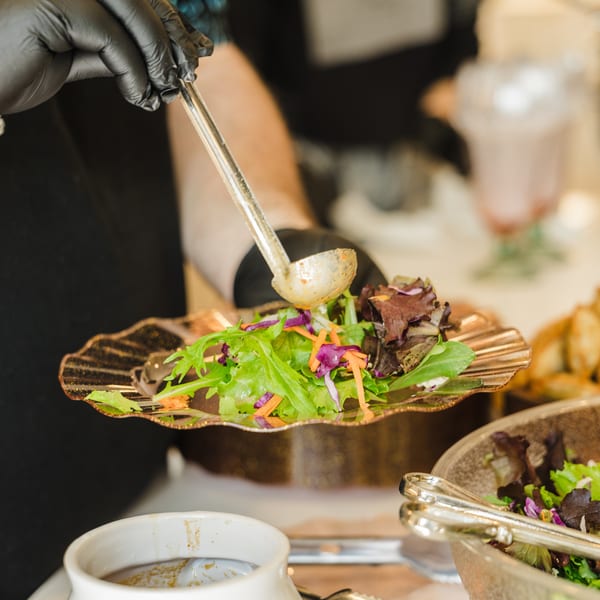 A gloved hand ladles dressing onto a fresh salad with mixed greens and shredded carrots.